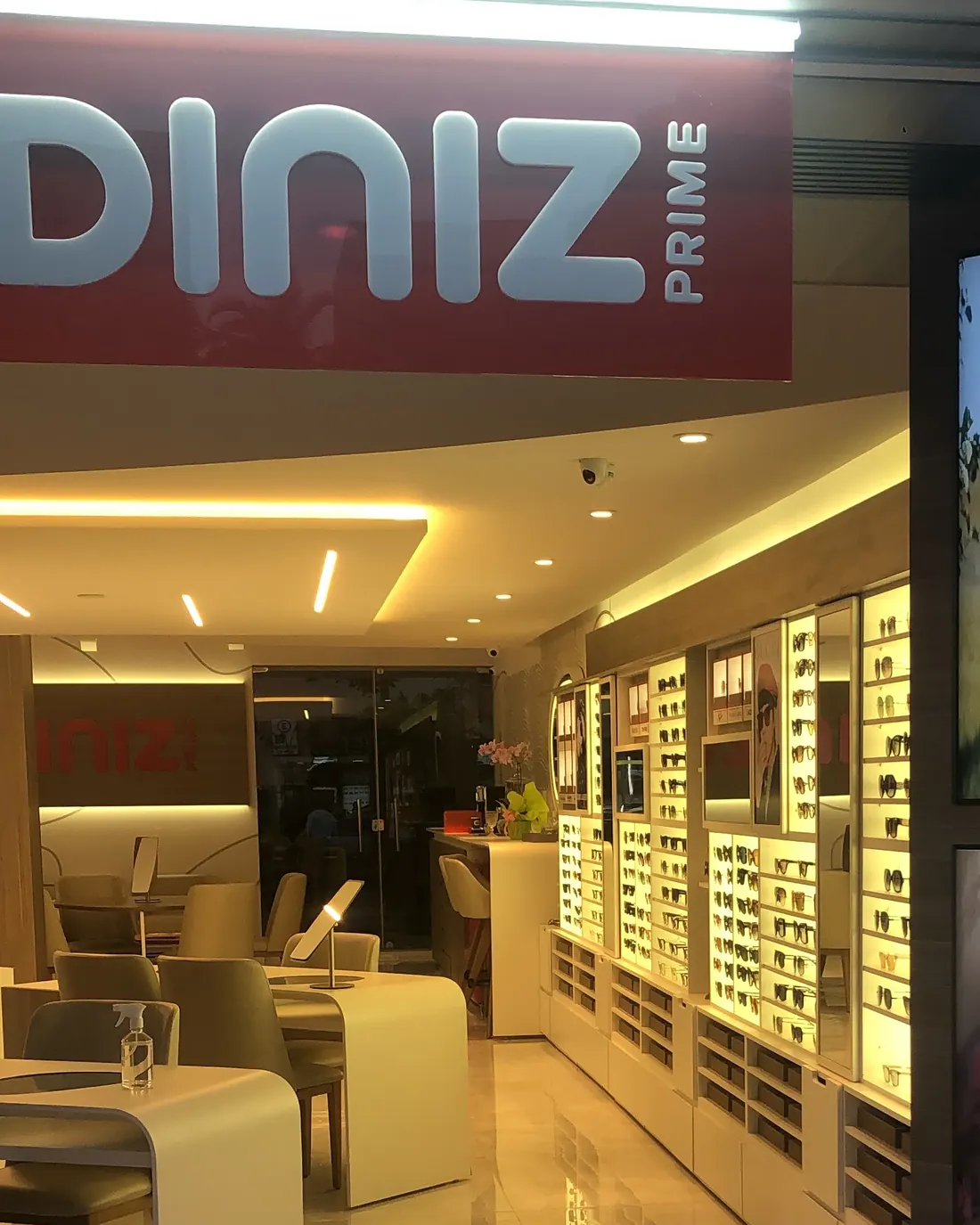 A entrada da Diniz Prime na 304 Sul ao anoitecer, com o letreiro, a parede de vitrines iluminadas e as mesas de atendimento.